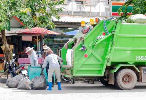 In this image, two sanitation workers are shown collecting and loading refuse into a bright green waste collection vehicle parked on a city street. One worker, wearing a grey uniform with blue rubber boots, is bending down to pick up a large black garbage bag while the other, also dressed in a grey uniform with blue gloves, stands near the vehicle's rear loading area, handling additional bags. The waste bins and bags are placed on the pavement beside the workers. In the background, a small open-front shop with a pink canopy and a red umbrella is visible, along with trees and various shop signage, indicating a lively urban environment. The waste collection vehicle is clean and modern, with visible equipment for loading rubbish. This scene reflects routine rubbish removal services frequently carried out in Maida Vale, part of London, close to W9 postcode, capturing the practical efforts involved in maintaining cleanliness in the area. The lighting appears natural, suggestive of daylight hours, with shadows cast on the ground and the vehicle.
