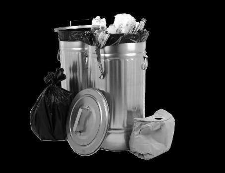 A large cylindrical metal outdoor rubbish bin with a closed lid, filled with mixed waste including crumpled paper, plastic, and cardboard, lined with a black trash bag that extends over the top of the bin. To the side, there is a tightly tied black plastic rubbish bag positioned on the ground, along with a white plastic bag that appears partially open. The scene is set against a plain black background, highlighting the metallic surface of the bin and the various recyclables and waste items surrounding it. The image showcases typical waste disposal contents, relevant to rubbish removal services in Maida Vale, emphasizing the need for professional clearance of household or office waste. The overall composition underlines the importance of proper rubbish management within local urban environments, such as London, while presenting a clear, detailed view of waste items ready for collection or disposal.