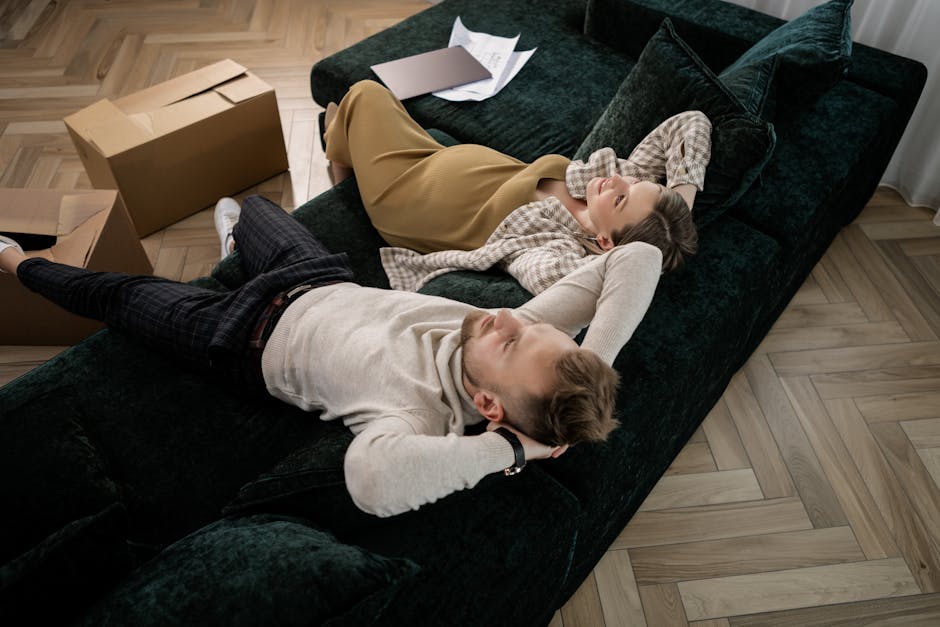 A three-seater upholstered sofa positioned outdoors against a brick wall, with the front of the sofa facing the camera. The fabric of the sofa is textured and appears worn, predominantly in a medium g
