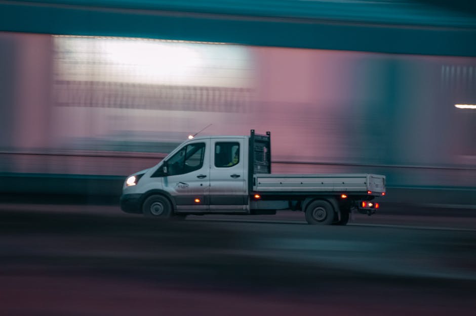 A white flatbed utility truck with black side mirrors and a black rear support structure is moving along a roadway, captured in motion with a blurred background indicating speed. The truck's body feat