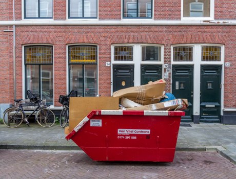 A large red skip container featuring a white warning label and contact number is situated directly in front of a row of three dark green front doors with white frames, attached to a red-brick building in an urban area. The skip is filled with various discarded items, including large cardboard boxes, flattened brown packing paper, and smaller miscellaneous debris, indicating ongoing rubbish removal activity. To the left of the skip, on the paved sidewalk, two bicycles are parked; one black, the other partially visible behind the skip, with a metal frame, baskets, and mudguards. The building façade above the doors shows windows with yellow curtains, and the pavement is made of interlocking bricks, typical of London street scenery. The overall scene depicts a typical rubbish clearance operation by Office Clearance Maidavale, serving local residents or businesses in the area near Maidavale, with a focus on waste removal from residential or commercial premises.