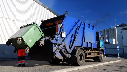 A large blue and black waste collection truck is positioned on a paved driveway next to a white industrial or commercial building under a bright blue sky. The truck's rear compactor is actively engaged in lifting or compacting a green plastic wheelie bin, which is fully emptied and placed at the edge of the vehicle's loading area. The green bin has four wheels and a lid that is open and tilted, revealing its interior. A worker dressed in a red high-visibility jacket and black trousers stands nearby, operating or supervising the process. The environment appears to be an industrial or business premises in Maidavale, with the truck's design and operational context emphasizing waste collection and rubbish removal services. The overall scene is well-lit by natural daylight, with clear weather conditions, and the surroundings include the building's exterior, concrete surface, and neat environment indicative of professional rubbish clearance activities. Office Clearance Maidavale provides rubbish removal solutions in this area, supporting efficient waste management for local businesses.