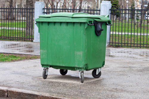 A green plastic wheeled rubbish bin with a closed lid, positioned on a paved surface in an outdoor area near a black metal fence with vertical bars. The bin appears to have a textured surface with some minor scuffs, and the wheels are visible at the bottom, facilitating mobility. In the background, there are trees with bare branches and a grassy area, suggesting a park or public space in a town with a postcode likely within the Maidavale region. The scene is evenly lit, indicating daytime, and the environment appears clean and well-maintained. This image relates to waste collection or rubbish removal services, possibly offered by Office Clearance Maidavale in the local area of Maidavale or nearby postcode, emphasizing the importance of proper rubbish disposal.