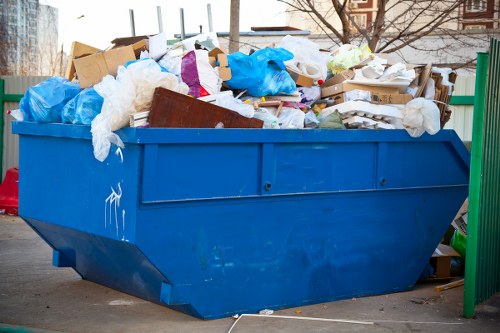 A large, rectangular blue rubbish skip filled with mixed waste materials, including white and brown paper, cardboard boxes, blue plastic bags, and various shredded packaging remnants, sits on a paved surface outdoors. The waste extends above the top edges of the skip, which is positioned near a green metal fence with vertical bars. In the background, faint outlines of buildings and trees suggest an urban or suburban area, potentially in Maidavale. The skip's surface appears weathered with some minor scratches, and the waste is loosely piled, indicating ongoing rubbish clearance work. The lighting suggests daylight, with natural light illuminating the scene clearly, capturing the textures of the paper, plastic, and cardboard among the waste. This image visually aligns with rubbish removal services provided by Office Clearance Maidavale, showcasing typical waste items encountered during office or property clearances in the local area.