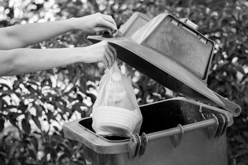 A person is shown disposing of a clear plastic bag filled with white household items into the top of a grey wheelie bin. The bin has a closed lid that is being lifted open, revealing a large black plastic interior. The person's hands are visible, guiding the bag into the bin, with a background of leafy shrubbery and outdoor foliage, indicating an outdoor setting likely on a driveway or pavement near a residential property. The scene captures a routine rubbish disposal process, commonly managed by companies such as Office Clearance Maidavale, highlighting waste collection and rubbish removal services in the local area. The bin appears to be made of durable plastic with a textured surface, and the lid is attached with hinges. The natural outdoor lighting creates a clear and neutral atmosphere, emphasizing the practicality of waste management in a typical residential environment in Maidavale.